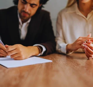 A couple sitting at a desk, signing legal documents. The man is holding a pen and signing papers, while the woman is holding a wedding ring in her hand. Who Qualifies for an Uncontested Divorce in Illinois