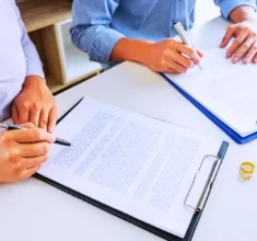 Two people signing divorce papers with wedding rings nearby.