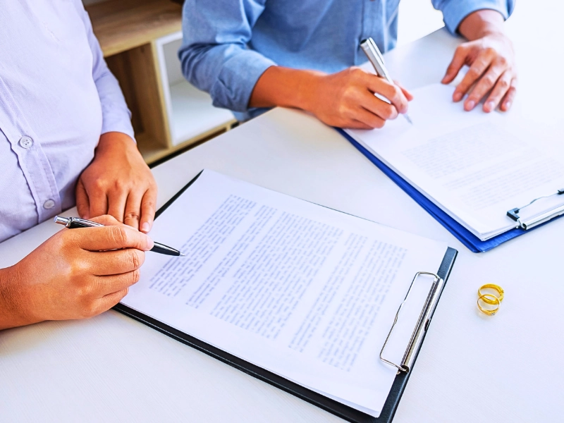 Two people signing divorce papers with wedding rings nearby.