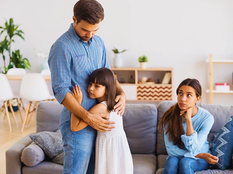Father hugging sad young daughter while mother sits on couch. uncontested divorce if you have children